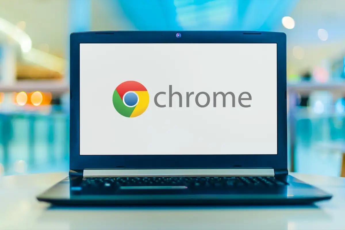 A laptop displays the Google Chrome logo and text on its screen; it rests on a table in a blurred, indoor shopping mall setting
